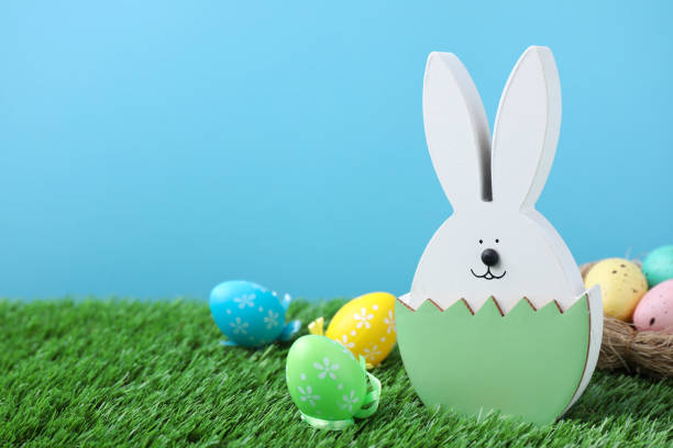 istock Easter bunny figure and dyed eggs on green grass against light blue background. Space for text 1334629367