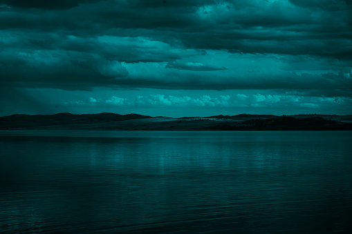 Blue green clouds over the sea. Toned seascape. Dark teal water and sky background with copy space for design. Rocky coastline on the horizon.