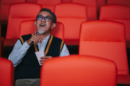 asian chinese active senior man enjoying his snack while watching movie in movie theater cinema alone