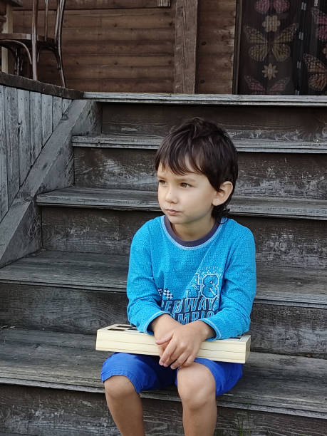 A five-year-old dark-haired boy sits on a wooden porch. He is holding a box with a chess set on his knees, his gaze is directed to the left. Close-up, outdoors, gray background. Bright blue shorts. un niño de cinco años de pelo oscuro se sienta en un porche de madera. está sosteniendo una caja con un juego de ajedrez sobre sus rodillas, su mirada se dirige hacia la izquierda. primer plano, al aire libre, fondo gris. pantalones cortos azules brilla