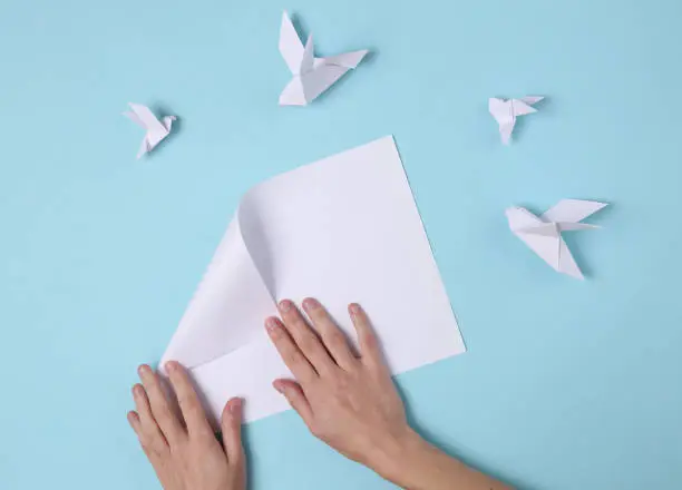 Female hands fold origami doves on a blue background. Top view