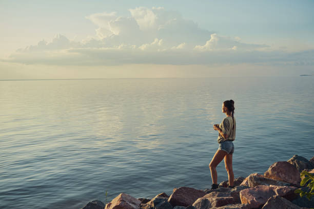 Female staying at the shoreline and staring at the sky and lake while drinking tea from steel cup