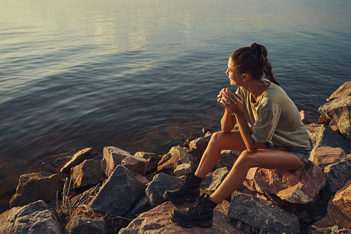 Woman turning her head and watching sun over the lake while drinking tea from steel mug on rocks at shore