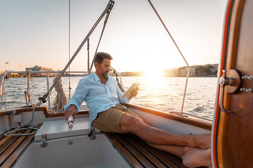 A man sitting alone on his sailboat, reading a paperback book in the sunset.