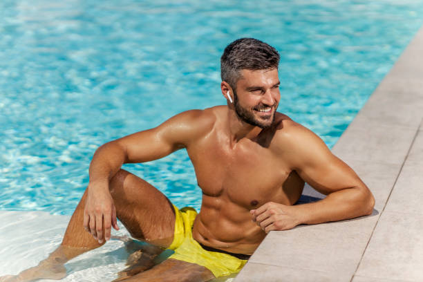 siente el sol y la música junto a la piscina. un hombre guapo se refresca en el agua de la piscina y escucha su canción favorita. nadar y tomar el sol, caluroso día de verano y disfrutar de los días de verano y descanso - swimming pool water people sitting fotografías e imágenes de stock