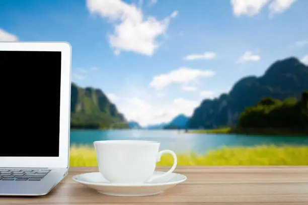 Photo of Coffee morning and laptop on wooden table on front of blurred background of the lake, mountain, meadow and blue sky among bright sunlight on a clear day.