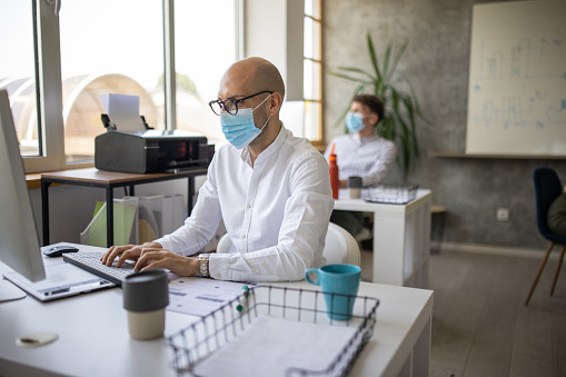 Businessman with protective face mask sitting at desk and working on computer in office