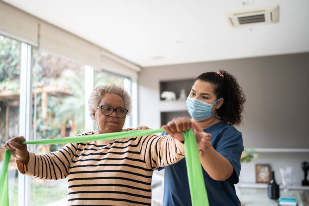 istock Senior woman doing exercise using resistance band with physical therapist support - wearing protective face mask 1334661779