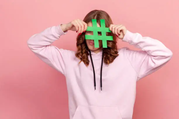 Photo of Portrait of curly haired teenage female in hoodie peeping through hashtag paper sign in hands, content for blog, popularity