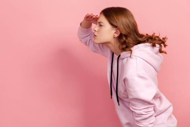 profil d’une adolescente curieuse aux cheveux bouclés en sweat à capuche tenant la main sur les yeux et regardant attentivement à gauche, essayant de voir quelque chose - far photos et images de collection