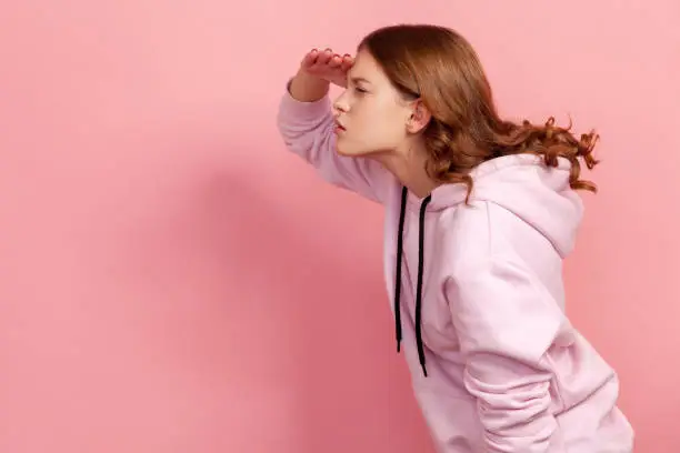 Profile of curious teenage girl with curly hair in hoodie holding hand over eyes and looking left attentively, trying to see something. Indoor studio shot isolated on pink background