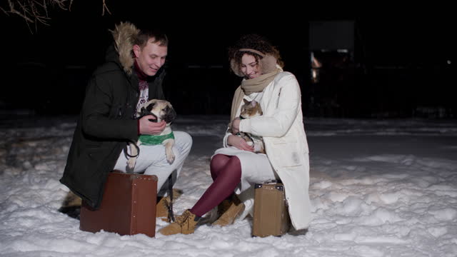 Man with dog and woman with cat sitting on travel suitcase at winter night. Romantic couple traveling together domestic pets at winter