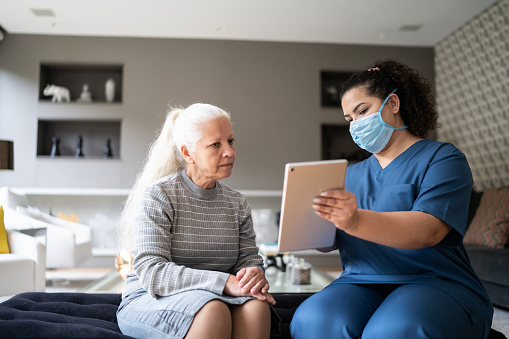 Nurse using digital tablet talking to senior patient at home during home visit - wearing protective face mask