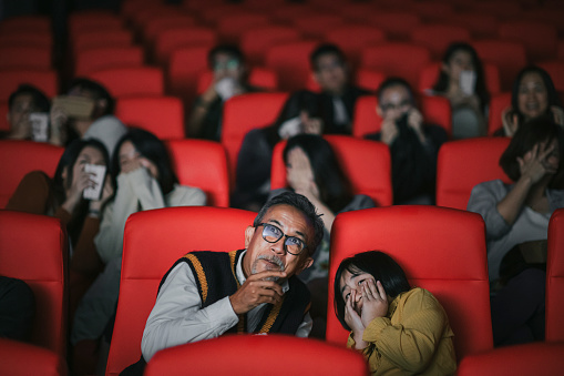 Asian chinese active senior man and his granddaughter watching horror scary movie in cinema movie theatre covering face