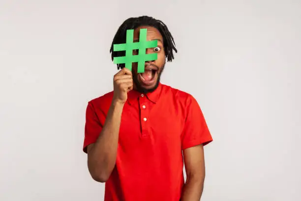 Photo of Guy with dreadlocks wearing red casual style T-shirt, covering face with social media hashtag symbol, recommending to follow trendy content, blog.