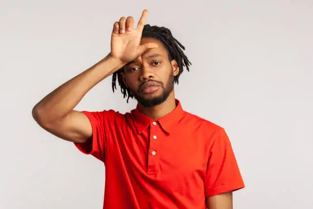 Handsome man with dreadlocks wearing red casual style T-shirt, showing looser gesture holding fingers near forehead, sad because of silly mistake. Indoor studio shot isolated on gray background.