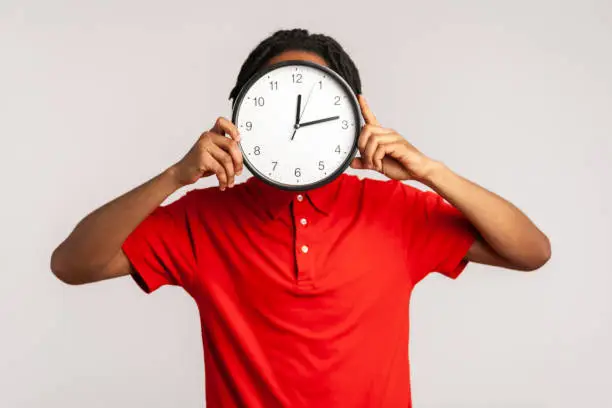 Photo of Unknown man with dreadlocks wearing red casual style T-shirt, hiding face behind big wall clock, time management, reminding of deadline.