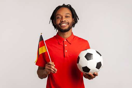 Man with toothy smile wearing red casual style T-shirt, holding flag of germany and soccer black and white classic ball and watching match. Indoor studio shot isolated on gray background.