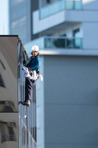 Image of a young skyscraper window washer wearing safety equipments and cleaning a glass curtain wall building facade