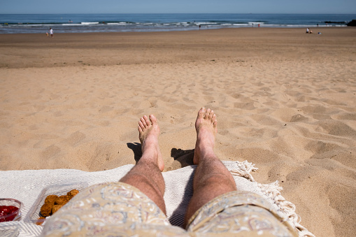 A point of view shot from an unrecognisable caucasian man wearing swim shorts on a beach in the North East of England. He is sitting on a picnic blanket and enjoying an array of snacks while enjoying the views of the sea.
