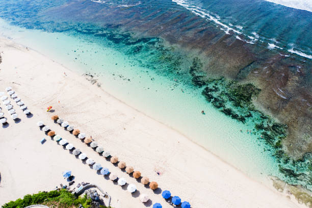 Beach scene - turquoise transparent ocean and blue beach umbrellas on the white sand beach. Bali, Melasti beach aerial view.