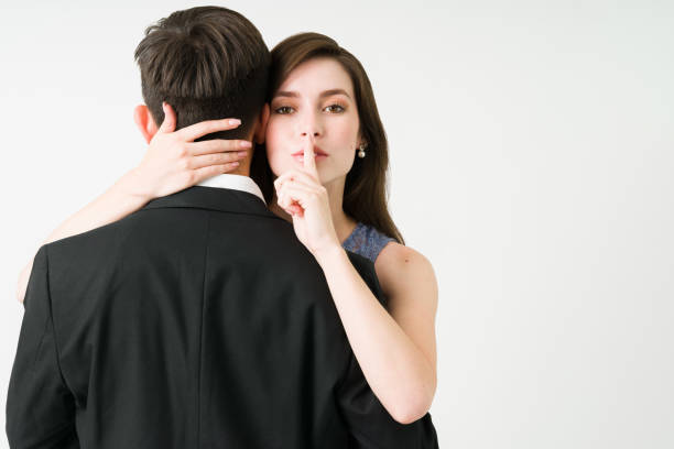 Mysterious woman cheating on her partner I have a secret. Portrait of a young girlfriend making eye contact and putting a finger on her mouth while hugging her boyfriend or husband whispering stock pictures, royalty-free photos & images