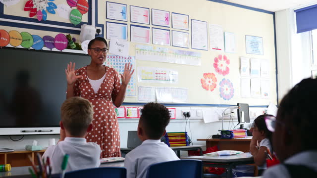 Pandangan sedang siswa sekolah dasar yang duduk di kelas yang diajarkan oleh seorang guru di Timur Laut Inggris. Para siswa menjawab pertanyaan dengan tangan mereka di udara sementara guru memberi isyarat kepada mereka.