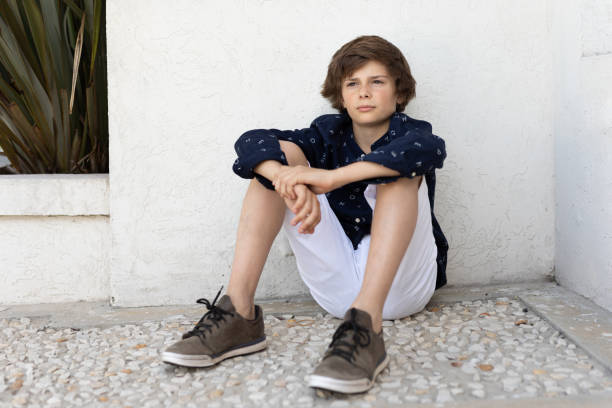 Vertical shot of preteen boy wearing blue sneakers and zip hoodie looking away while sitting on floor posing in studio against neutral background, copy space