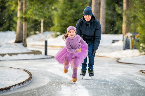 little girl in a pink sweater and a fluffy skirt on sunny winter day rides with her dad on an open skating rink in park
