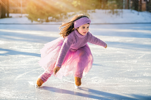 little girl in a pink sweater and a full skirt rides on a sunny winter day on an outdoor ice rink in the park