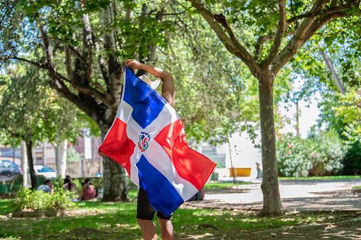 Handsome latino man holding the Dominican Republic flag on an out of focus outdoors background. Selective focus: Diversity concept.