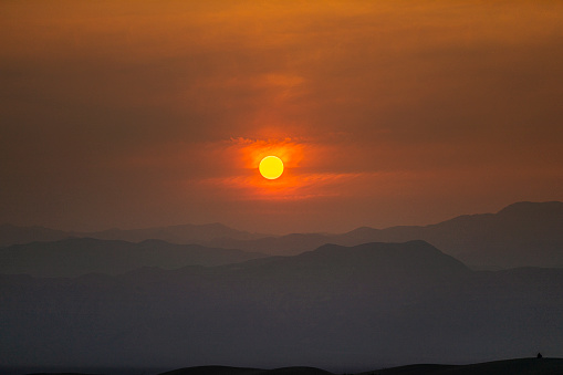 Bright orange sun setting behind mountain range in desert with smoke orange sky from forest fires