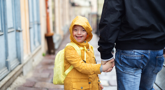 A happy down syndrome boy with unrecognizable father outdoors on a walk in rain, looking at camera.