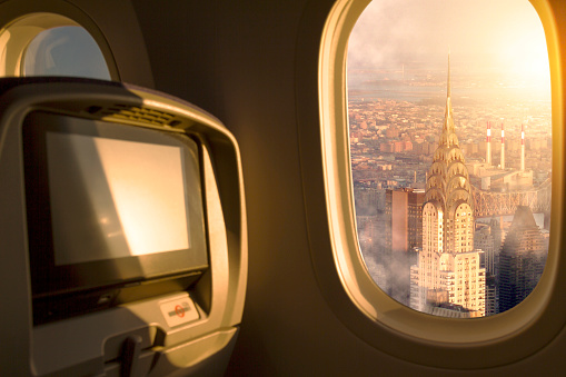 New York City, USA at sunset / sunrise sky aerial view from porthole window airplane economic seat after take off from airport, on back Chrysler Building, Midtown Manhattan, Queens and Bronx District. Travel concept. Plane interior.