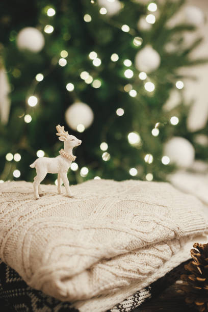 istock Cozy knitted sweaters with reindeer toy and pine cone on rustic wood on background of christmas tree lights. Pile of stylish sweaters in festive decorated scandinavian room.  Winter holidays 1334711023