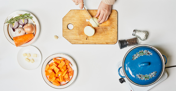 Woman hand cutting onions. Preparing pumpkin soup. Ingredients of pumpkin soup on white background. Top view. Organic fresh pumpkin, onion, rosemary, garlic and salt with portable electric stove