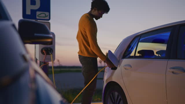 Slow motion dolly shot of a young man uses his smartphone while he waits for his car to be charged on the parking lot at dusk. Blue light illuminates the interior of the car and the electric vehicle charging station.