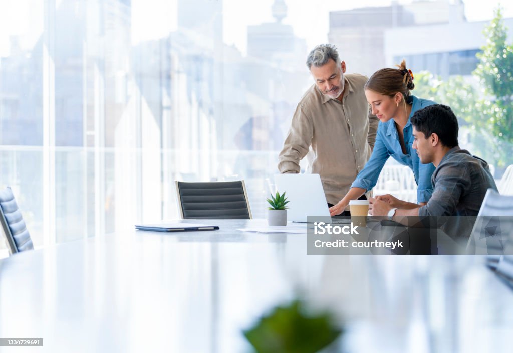Group of business people working. Group of business people working. The team are standing and sitting at a board room table looking at a laptop computer. All dressed in casual business clothing with a window behind them Business Stock Photo