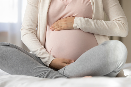 Late pregnant woman sitting in beddings, touching, caressing baby bump covered pink cloth. Expecting mom relaxing at home, enjoying leisure time in pregnancy, holding, embracing tummy. Cropped shot.