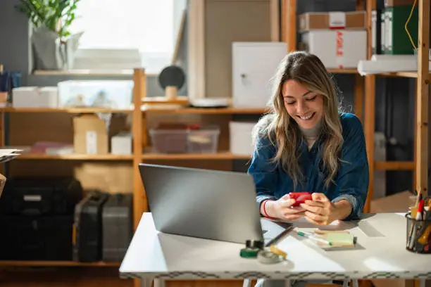 Photo of Boosting Productivity: Young Happy Caucasian Female Entrepreneur Sitting at her Storage Facility, Multitasking on her Computer and Smartphone ( Horizontal)