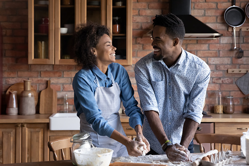 Overjoyed millennial Africa American couple enjoy family weekend cooking together work with dough in kitchen. Smiling young ethnic man and woman bake pastry or cookies at home. Hobby concept.