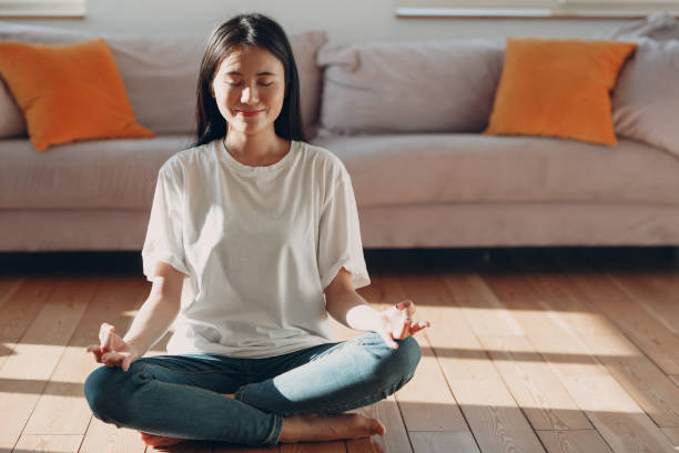 Asian woman doing yoga and zen like meditation lotus pose in casual wear at indoor living room apartment with natural sun light stock photo