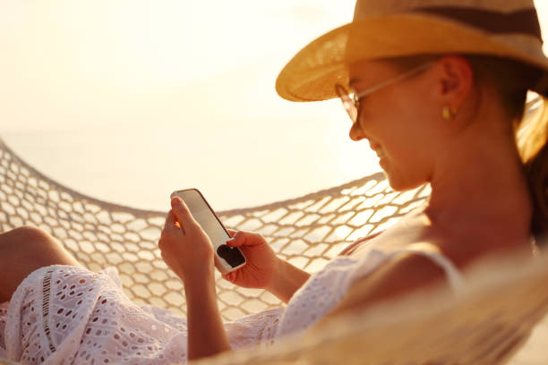 istock Working at sunset. Young happy female using smartphone while lying in the hammock on the beach 1334750829
