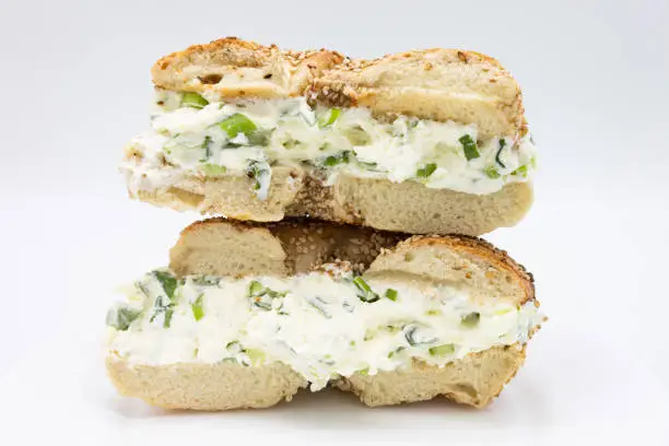 A cut in half and stacked New York style sesame seed bagel filled with scallion cream cheese with a white background