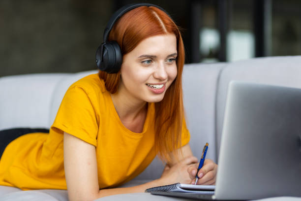 istock Smiling happy young woman with red hair watching a webinar using laptop computer lying on the couch at home. Online education, learning at home concept 1334770361