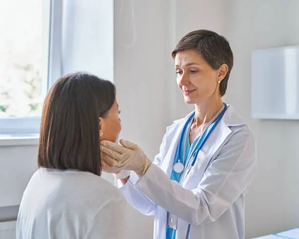 Friendly middle-aged woman doctor wearing gloves checking sore throat or thyroid glands, touching neck of young african female patient visiting clinic office. Thyroid cancer prevention concept