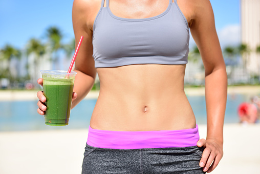 Healthy lifestyle fitness woman drinking green vegetable smoothie juice after running exercise. Close up of smoothie and stomach. Healthy lifestyle concept with fit female model outside on beach.