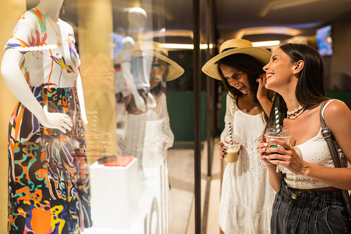 Two young women are having fun window shopping with coffee to go on a summer evening