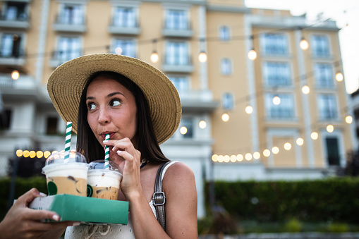 A pretty young woman is drinking iced coffee from a disposable cup through a straw outdoors in summer