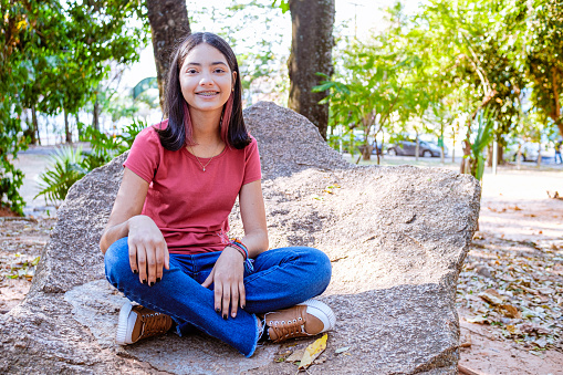 Girl sitting on stone during park walk, smiling at camera. Adolescent portraits.